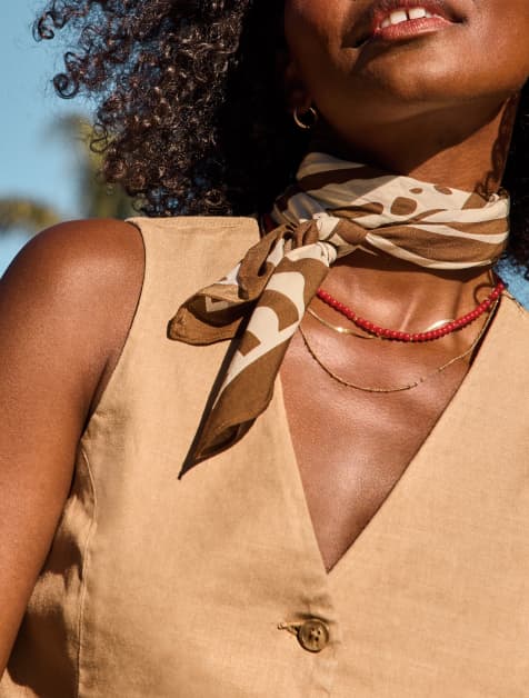 Image of woman wearing a scarf tied around her neck with a stack of necklaces and tan vest