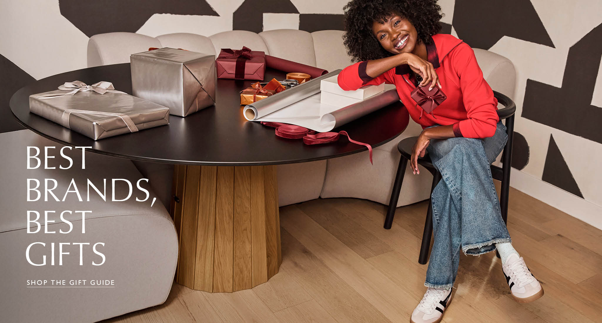 Woman sitting at a table wrapping presents wearing a red sweatshirt with wide leg jeans and sneakers. Text over the image reads “Best Brands, Best Gifts, SHOP THE GIFT GUIDE”