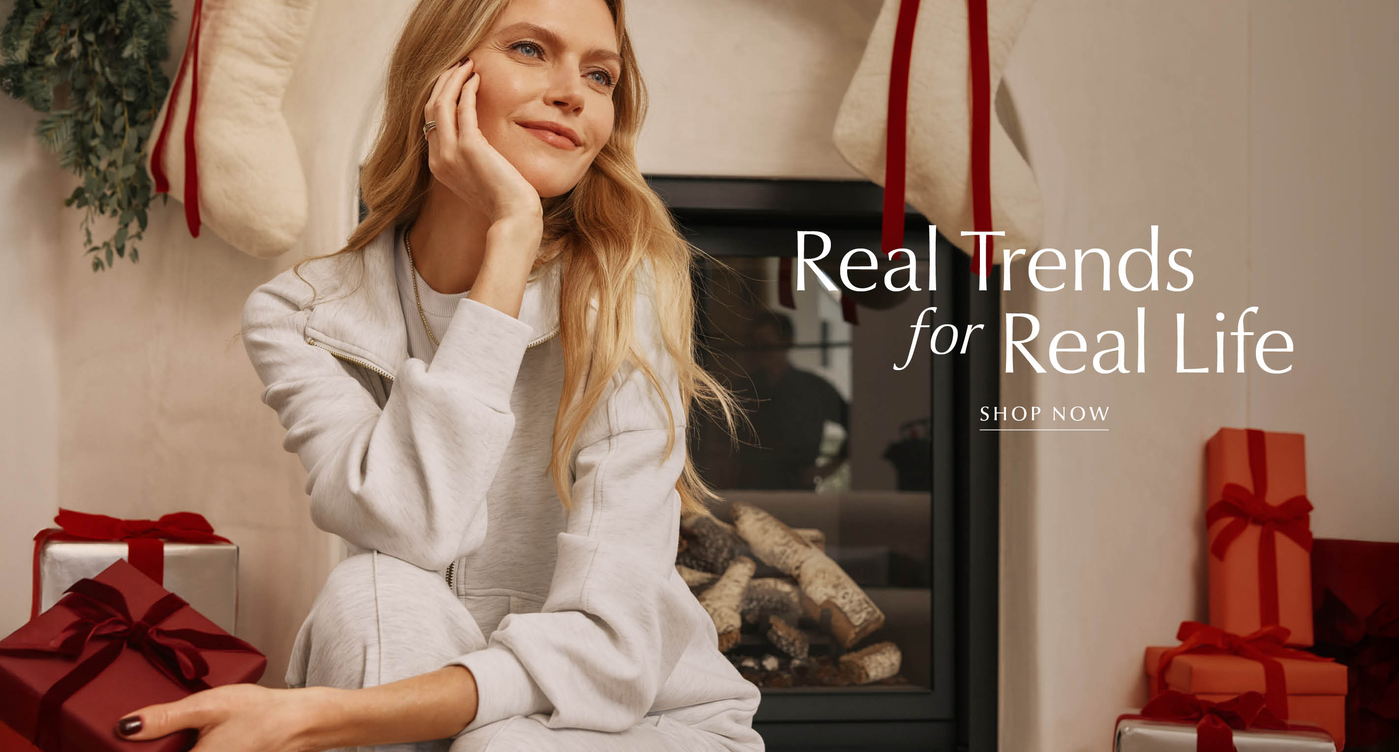 Woman sits by a fireplace wearing a grey zip up sweatshirt with matching wide leg pants and sherpa slippers, text over image reads: Real Trends for Real Life, Shop Now.