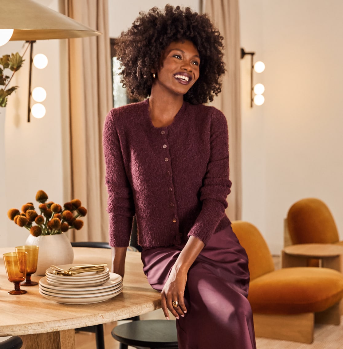 Woman leaning on a table wearing a burgundy cardigan sweater with a matching burgundy satin midi skirt. 