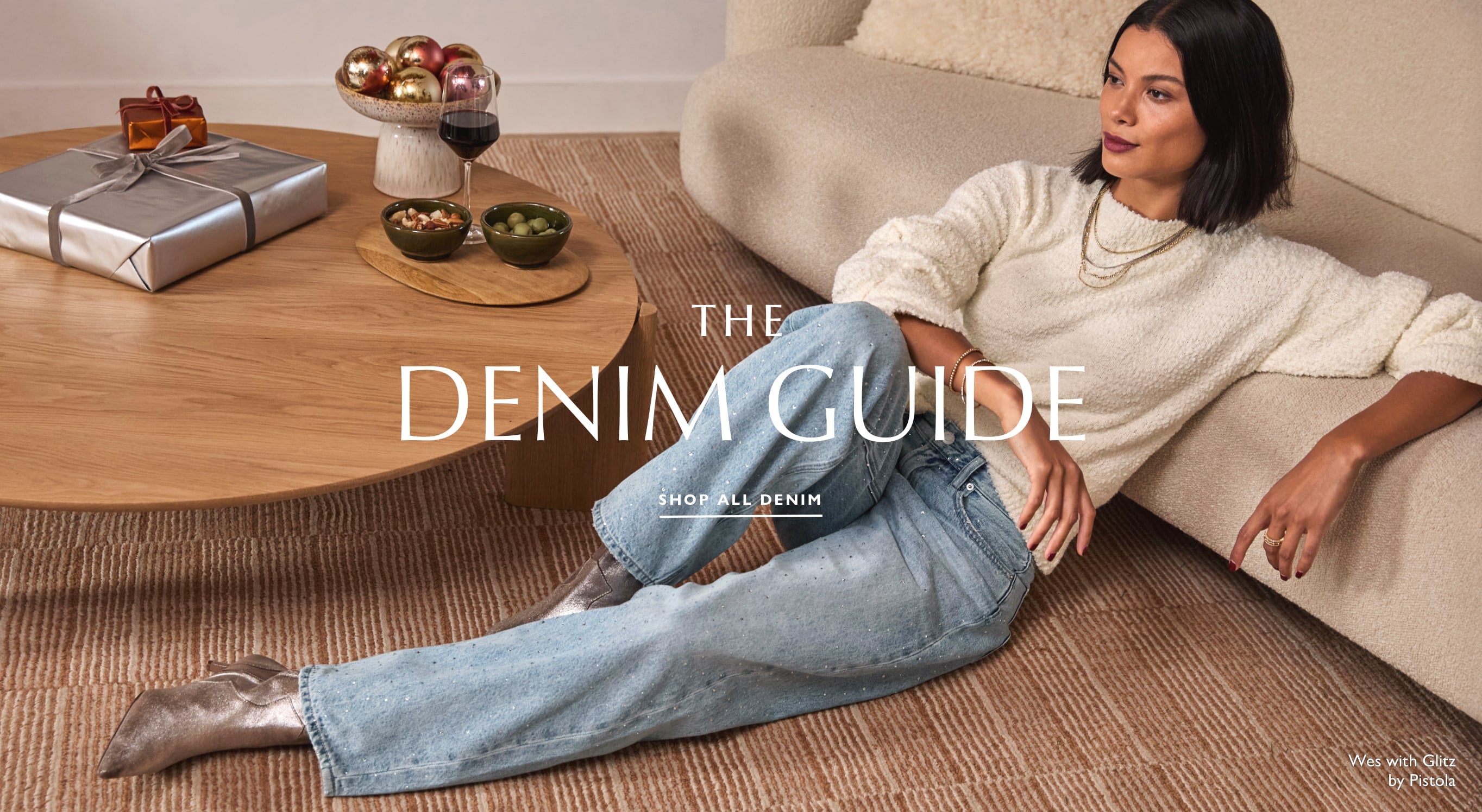 Woman lounging on a rug wears an ivory sweater with sparkly jeans called the "Wes with Glitz" jean by Pistola. Text over image reads: "The Denim Guide, Shop all Denim". 