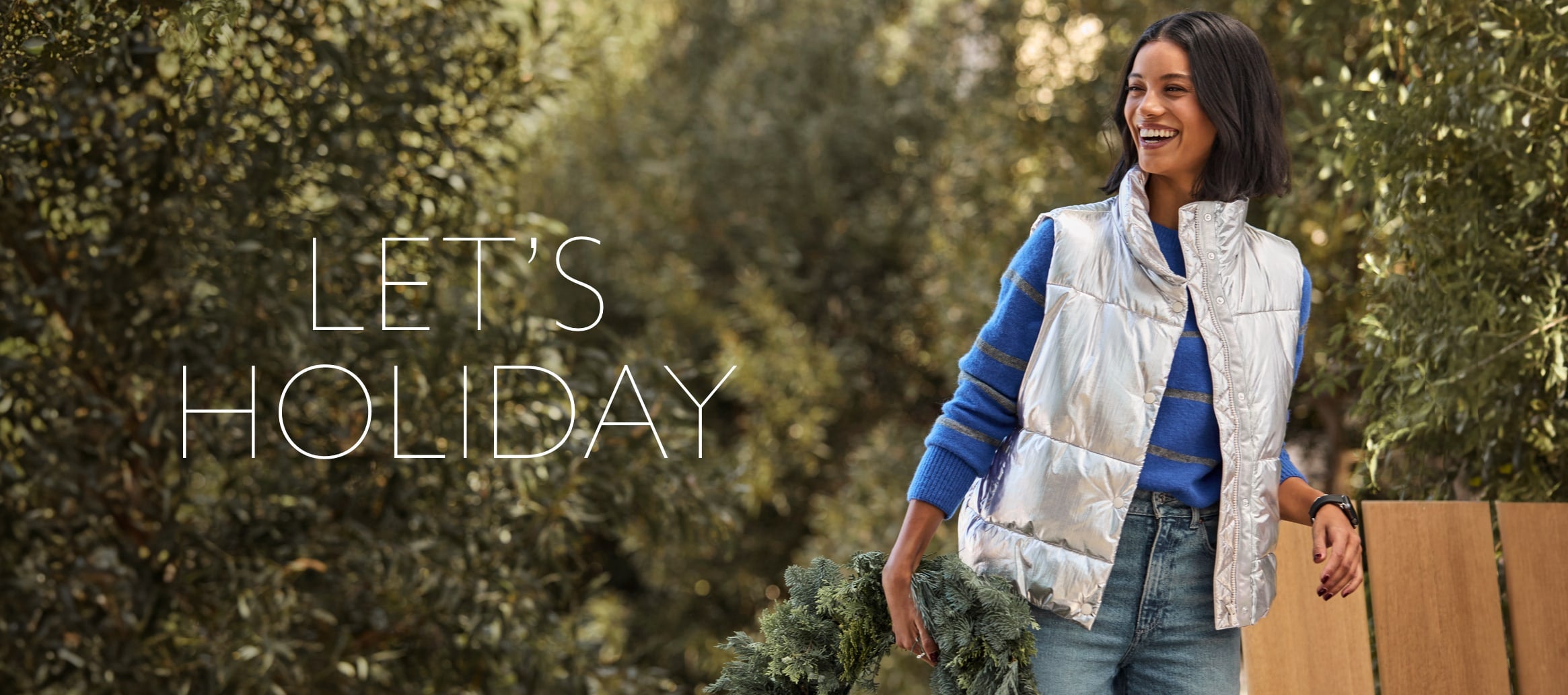 Woman carrying a wreath wears a blue crew neck sweater with grey stripes layered with a metallic silver puffer vest and jeans. Text over image reads Let's Holiday