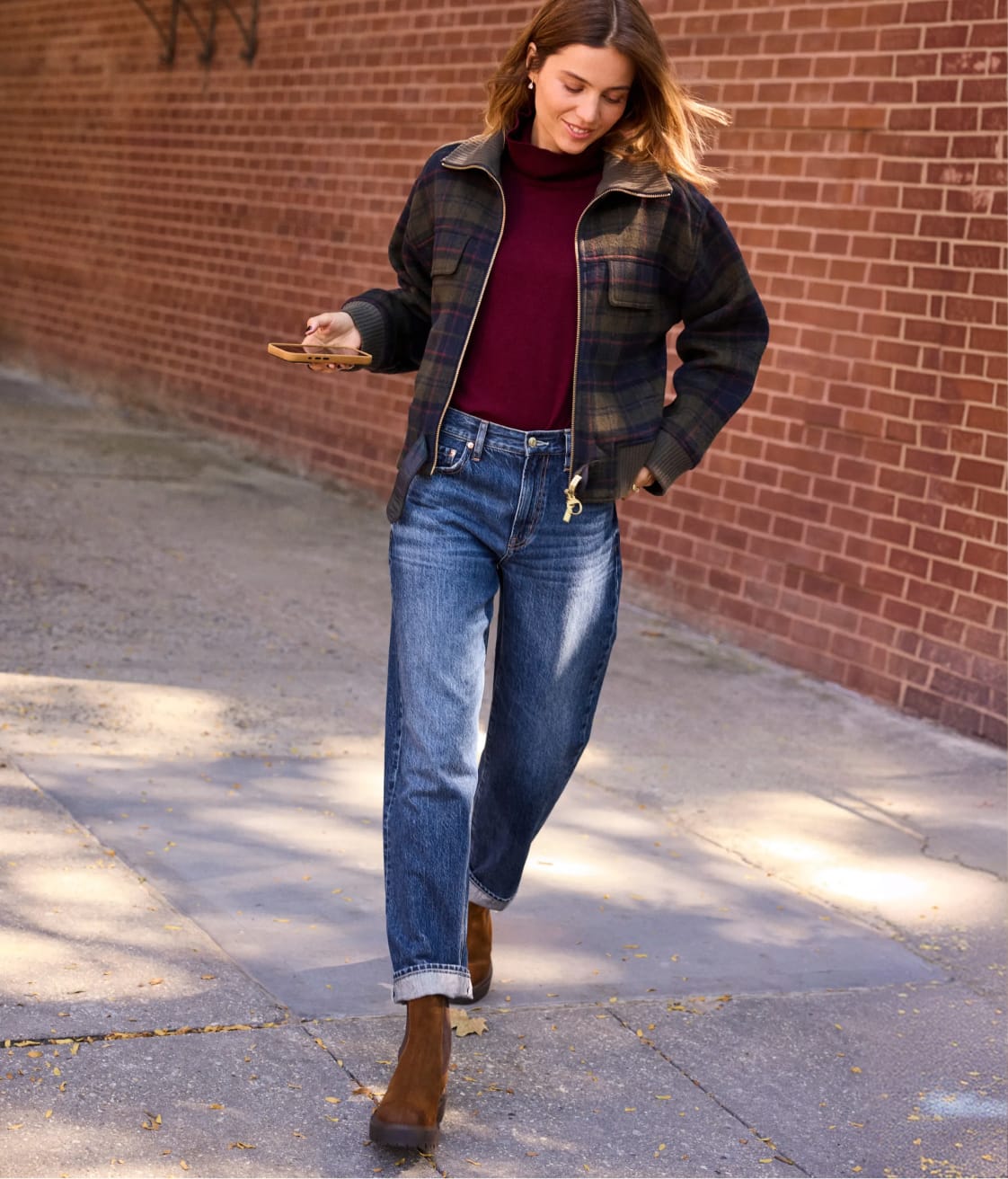 Woman wearing plaid cropped jacket over a burgundy sweater with barrel jeans and brown boots.